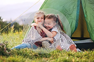 Two little hugging girls sisters sitting on the green grass next camp tent entrance, cheerfully smiling. Careless childhood,