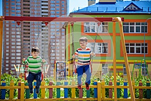 Two little kid boys having fun with swing on outdoor playground. Children, best friends and siblings swinging on warm sunny spring