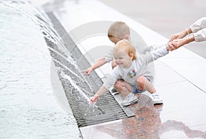 Two little kids playing in the city square fountain. Children hold mummy by a hand.