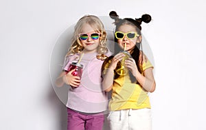 Two little models in sunglasses, colorful casual clothes. Holding cocktail bottles, showing tongues, posing isolated on white