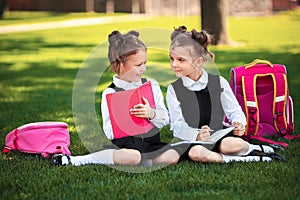 Two little school girls with pink backpack sitting on grass after lessons and read book or study lessons, thinking ideas