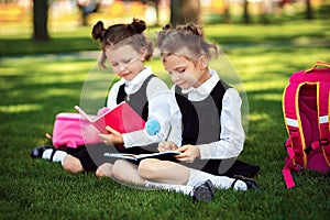 Two little school girls with pink backpack sitting on grass after lessons and read book or study lessons, thinking ideas