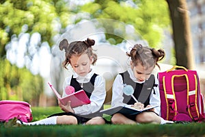 Two little school girls with pink backpack sitting on grass after lessons and read book or study lessons, thinking ideas