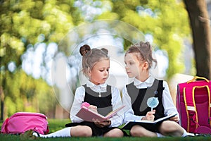 Two little school girls with pink backpack sitting on grass after lessons and read book or study lessons, thinking ideas