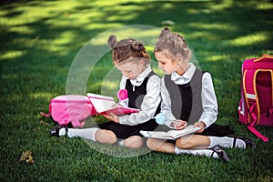 Two little school girls with pink backpack sitting on grass after lessons and read book or study lessons, thinking ideas