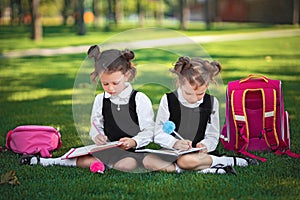 Two little school girls with pink backpack sitting on grass after lessons and read book or study lessons, thinking ideas