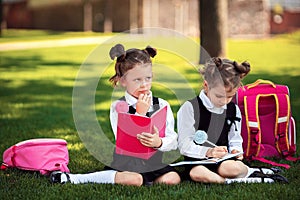 Two Little school girls with pink backpack sitting on grass after lessons and thinking ideas, read book and study