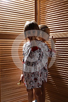 Two little sisters dressed in the pajamas are hiding in the closet with wooden doors