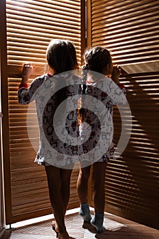 Two little sisters dressed in the pajamas are hiding in the closet with wooden doors