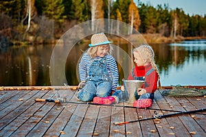 Two little sisters or friends sit with fishing rods on a wooden pier. They caught a fish and put it in a bucket. They are happy