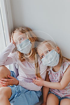 Two little sisters with masks on their faces look out of the window during the quarantine hugging each other