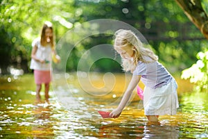 Two little sisters playing with paper boats by a river on warm and sunny summer day. Children having fun by the water.