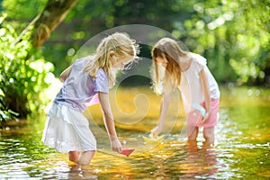 Two little sisters playing with paper boats by a river on warm and sunny summer day. Children having fun by the water.