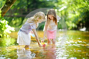 Two little sisters playing with paper boats by a river on warm and sunny summer day. Children having fun by the water.