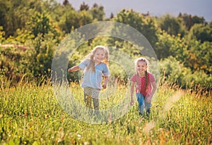 Two little sisters running and laughing in the summer high green grass meadow with a bright sunset light background. Careless