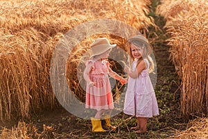 Two little sisters in straw hats and pink dresses are running around in a wheat field, having fun