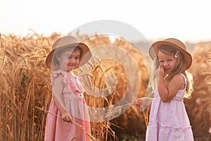 Two little sisters in straw hats and pink dresses are running around in a wheat field, having fun