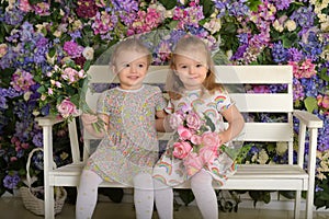 Little twin girls in the garden on a bench against the background of a floral wall with bouquets of flowers in their hands