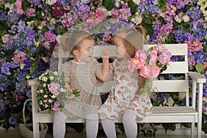 Little twin girls in the garden on a bench against the background of a floral wall with bouquets of flowers in their hands