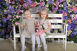 Little twin girls in the garden on a bench against the background of a floral wall with bouquets of flowers in their hands