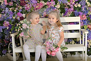 Little twin girls in the garden on a bench against the background of a floral wall with bouquets of flowers in their hands