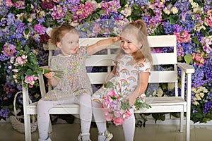 Little twin girls in the garden on a bench against the background of a floral wall with bouquets of flowers in their hands