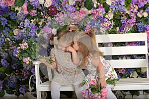 Little twin girls in the garden on a bench against the background of a floral wall with bouquets of flowers in their hands