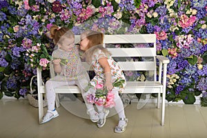 Little twin girls in the garden on a bench against the background of a floral wall with bouquets of flowers in their hands