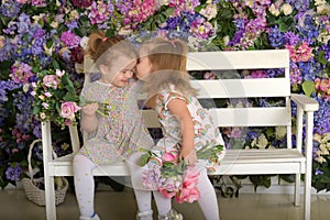 Little twin girls in the garden on a bench against the background of a floral wall with bouquets of flowers in their hands