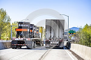 Two loaded big rig semi trucks with different semi trailers running side by side on the narrow two line highway intersection at