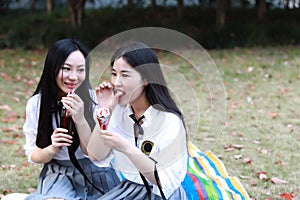 Two lovely Asian Chinese pretty girls wear student suit in school best friends smile laugh drinking cola eating icecream on grass