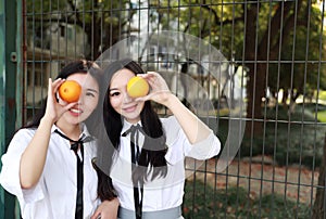 Two lovely Asian Chinese pretty girls wear student suit in school best friends smile laugh hold orange fruit on the playground