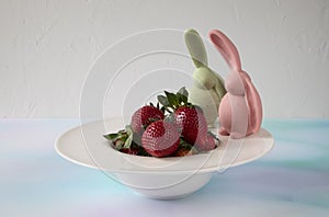 Two lovely chocolate bunnies sit on the edge of a plate. The plate contains beautiful, fresh and ripe strawberries, mesmerizing