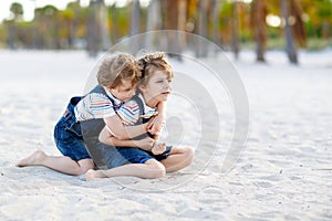 Two lovely little brothers sitting on sand on tropical beach, happy best friends, kids boys playing, friendship concept