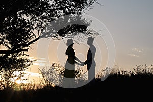 Two lovers girl and a man stand under a beautiful tree in the rays of the sunset in the evening of summer. Beautiful romantic