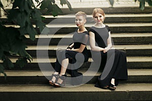 Two luxury gorgeous woman in black dress posing, sitting on old stairs in soft light in city, classic gothic lady outfit