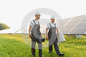 Two male electrician workers walking in between long rows of photovoltaic solar panels and talking about installation of