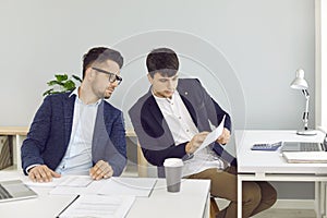 Two male employees sitting at office desks and looking at some business documents