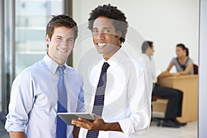 Two Male Executives Using Tablet Computer With Office Meeting In Background