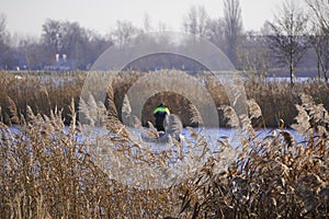 Two male fishermen in warm clothes on a boat, winter fishing on a river or lake. Photo through dry, brown reeds or grass