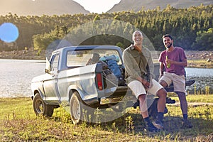 Two Male Friends With Backpacks In Pick Up Truck On Road Trip By Lake Drinking Beer