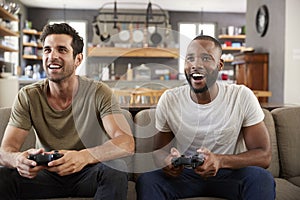 Two Male Friends Sitting On Sofa In Lounge Playing Video Game