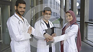 Two male handsome doctors and their pretty female colleague in hijab, stacking hands