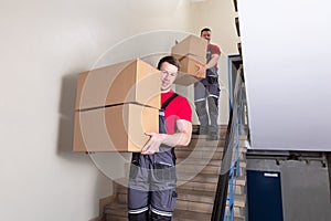 Two Male Movers Walking Downward With Boxes On Staircase