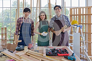 Two male and one female asian adult carpenters holding building tools and stand smiling together behind working desk full of