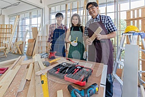 Two male and one female asian adult carpenters holding building tools and stand smiling together behind working desk full of