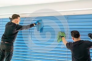 Two male professional cleaners cleaning blue blinds on a large window front with micro fiber cloths