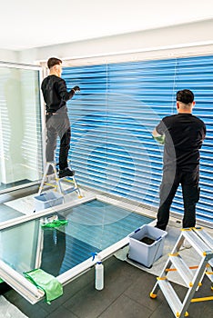 Two male professional cleaners cleaning blue blinds on a large window front with micro fiber cloths