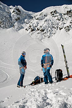 two male skiers in ski suits and helmets stand against the backdrop of a snow-covered mountain slope.