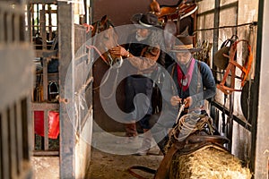 Two man with cowboy costume stay in horse stable that one check the bullet of short gun and other one take care horse in the back